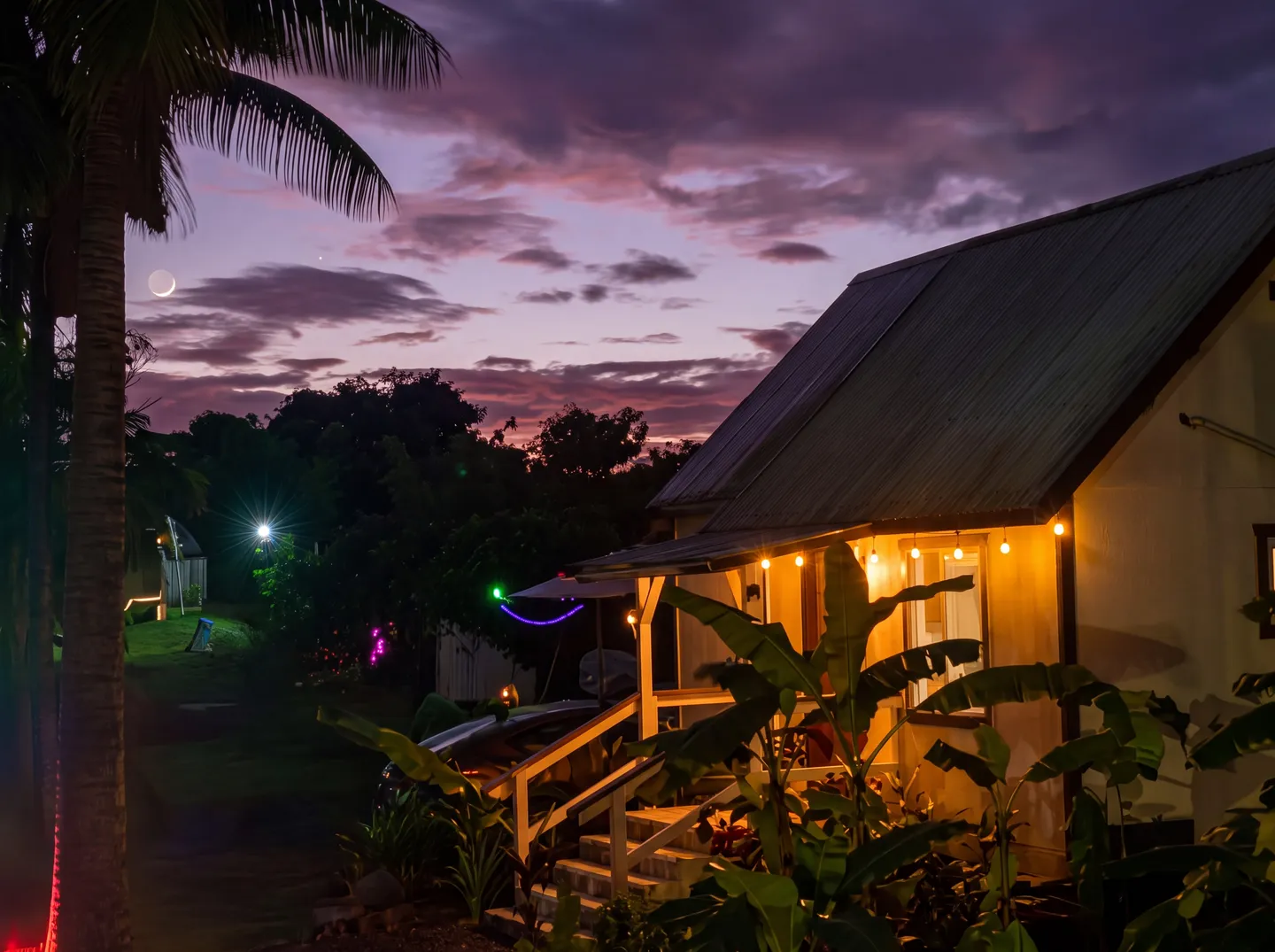 Exterior of a cottage at dusk with string lights illuminating the porch beneath a purple and pink sunset sky with a crescent moon.
