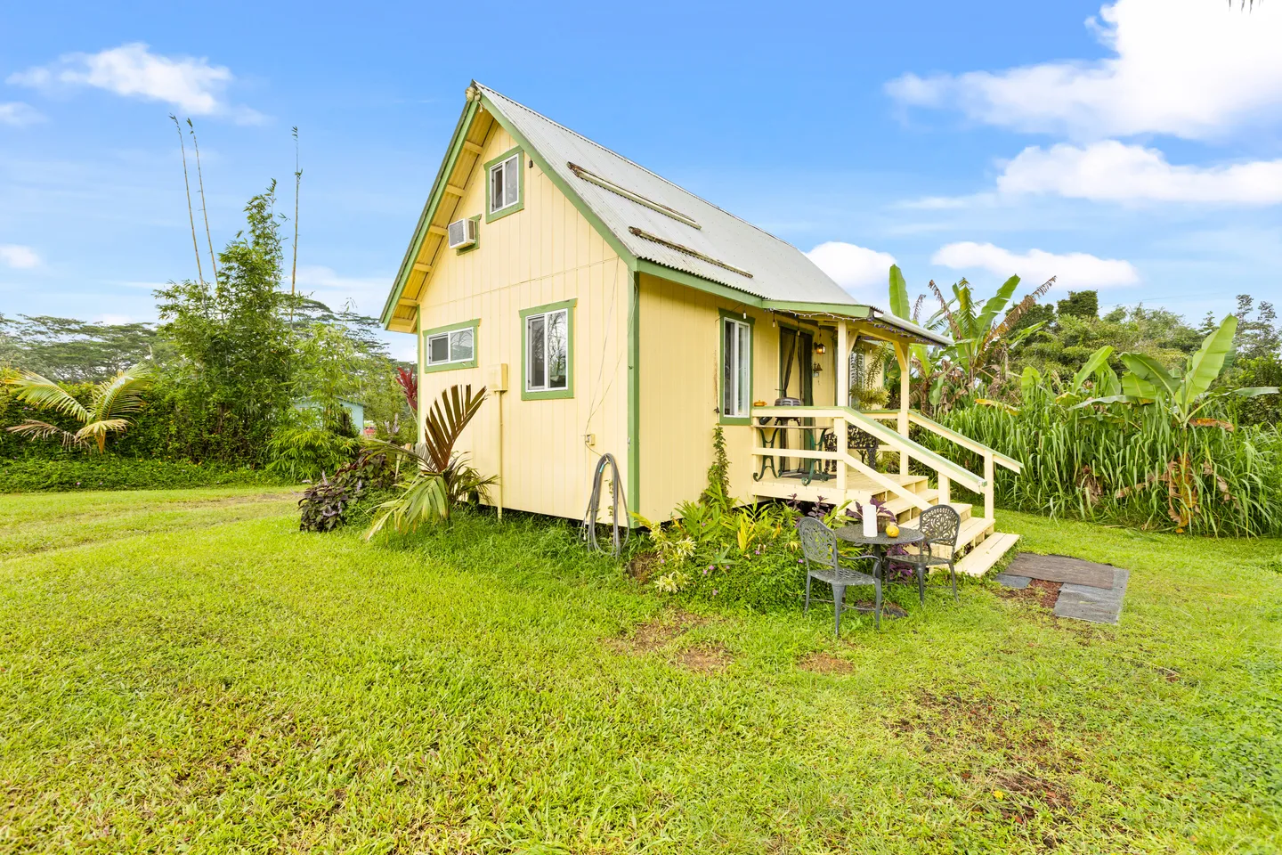 Exterior of a small yellow cabin with green trim, a metal roof, and a small front porch surrounded by a grassy lawn and tropical foliage.