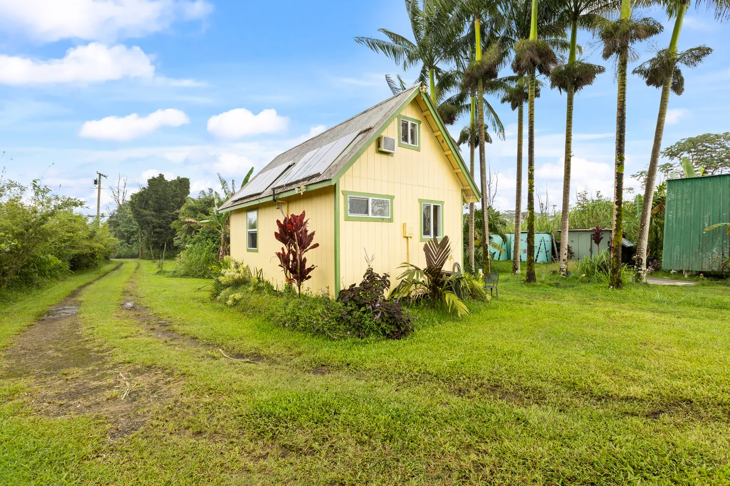 Exterior of a small yellow cabin with green trim, solar panels on the roof, surrounded by tropical foliage and palm trees under a blue sky.
