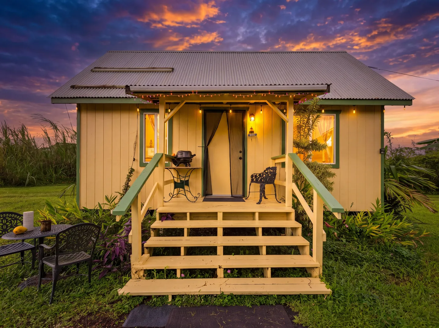 Exterior of a yellow cottage with a covered front porch and stairs set against a colorful sunset sky.