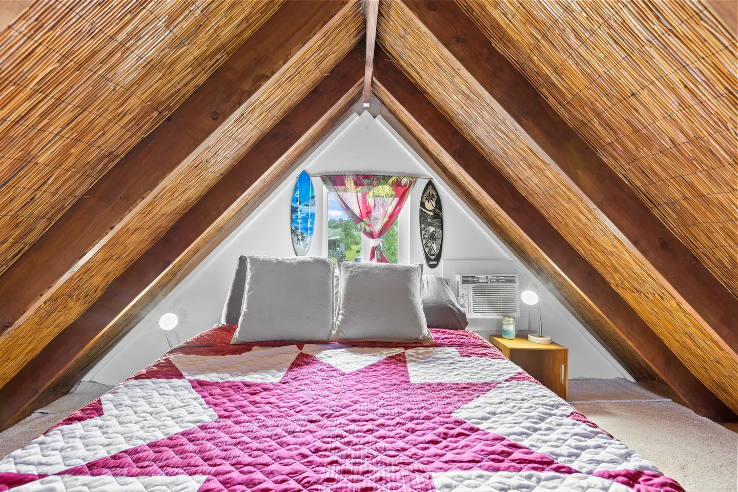 A-frame loft bedroom with a pitched thatched ceiling, exposed beams, and a bed topped with a pink and white star-patterned quilt.