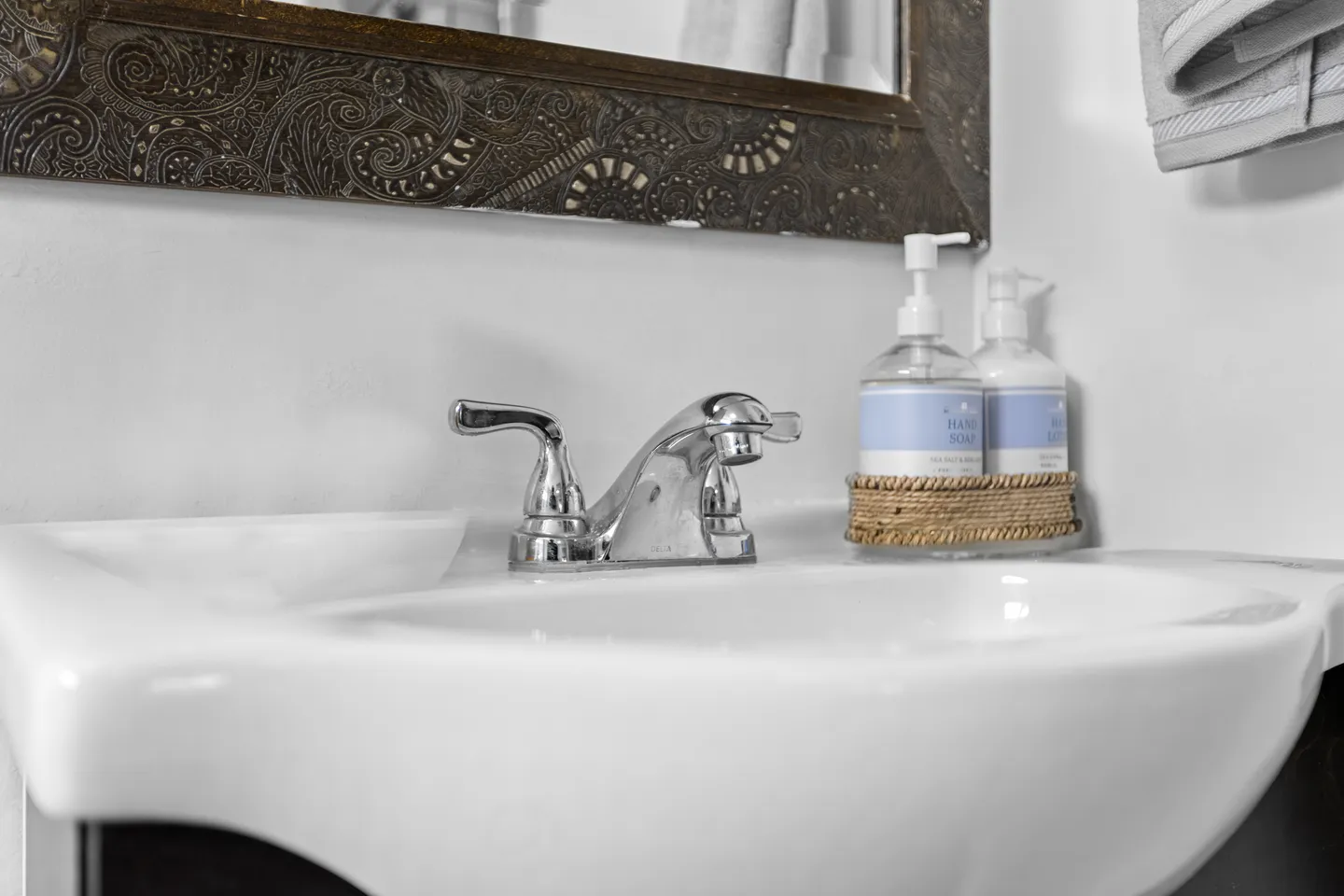 Close-up of a white bathroom sink with a chrome faucet, ornate mirror frame, and soap dispensers.