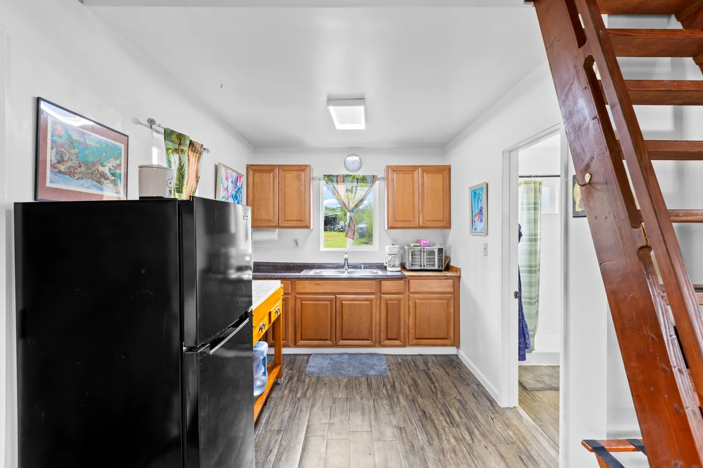 Kitchen with wooden cabinetry, a dark refrigerator, a sink under a window, and wooden stairs on the right.