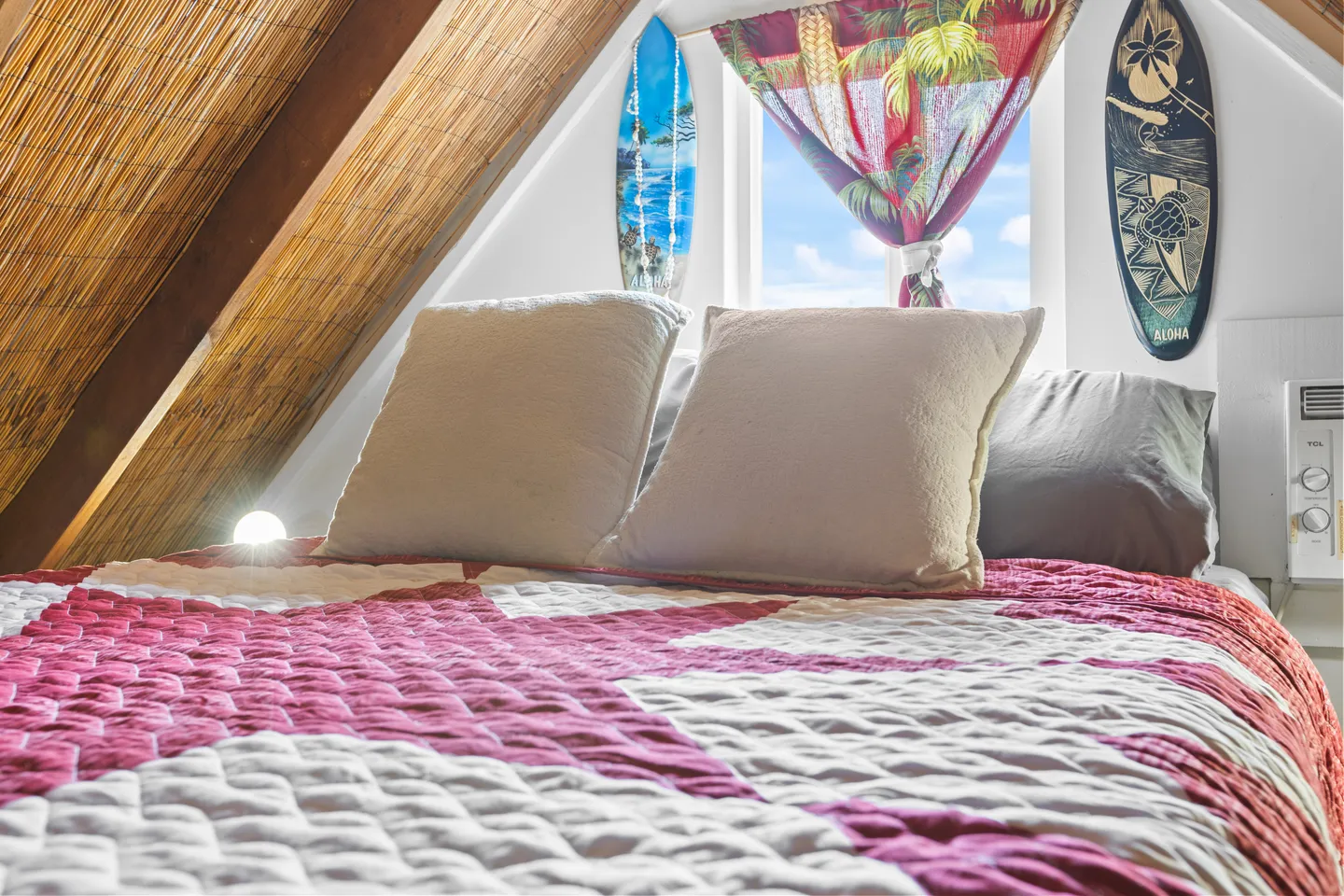 A close-up view of a bed with a pink and white quilt beneath an angled thatched-style ceiling, flanked by decorative mini surfboards and a window.