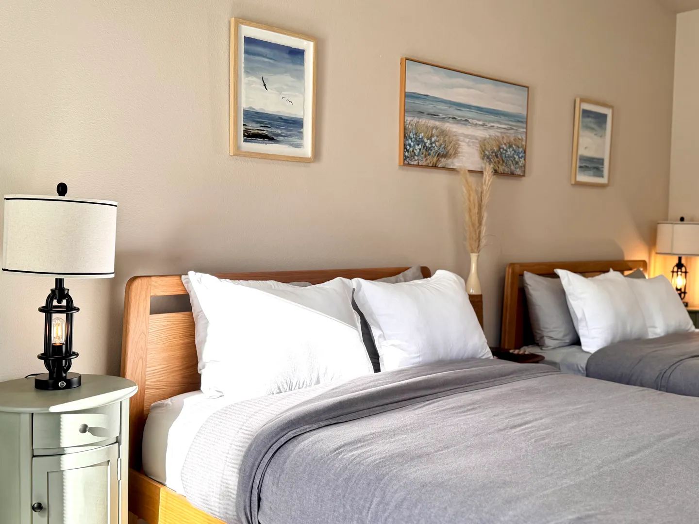 Bedroom featuring two wooden beds with grey blankets and white pillows, coastal artwork on the wall, and bedside tables with lamps.