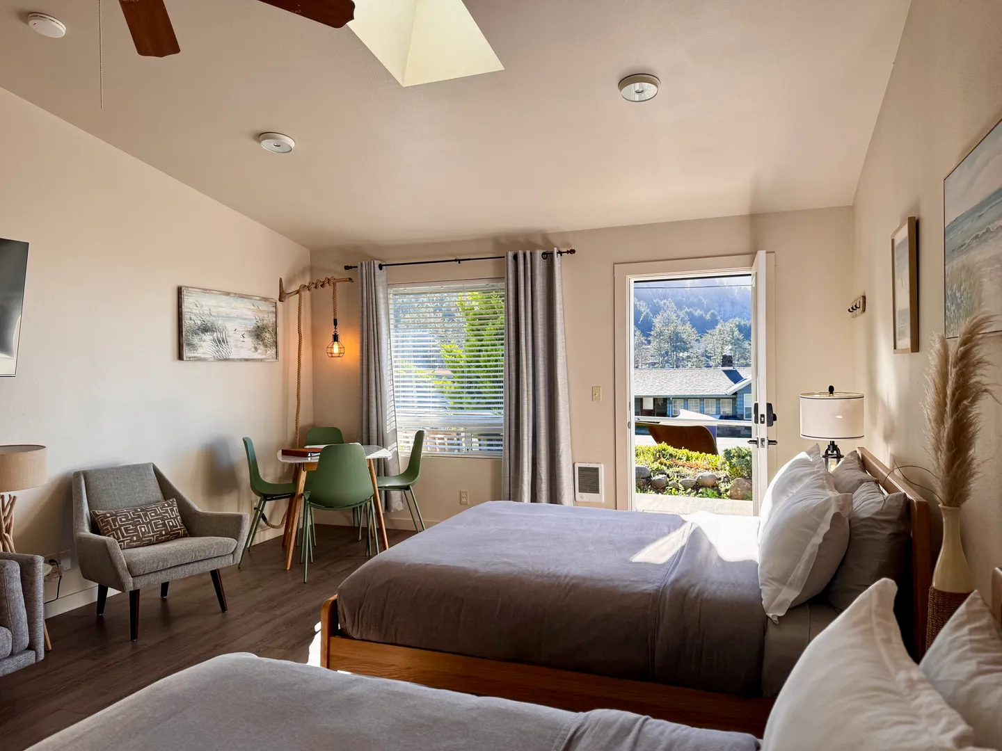 Bedroom featuring two beds, an armchair, a small dining table with chairs, a window, and an open exterior door looking outside.