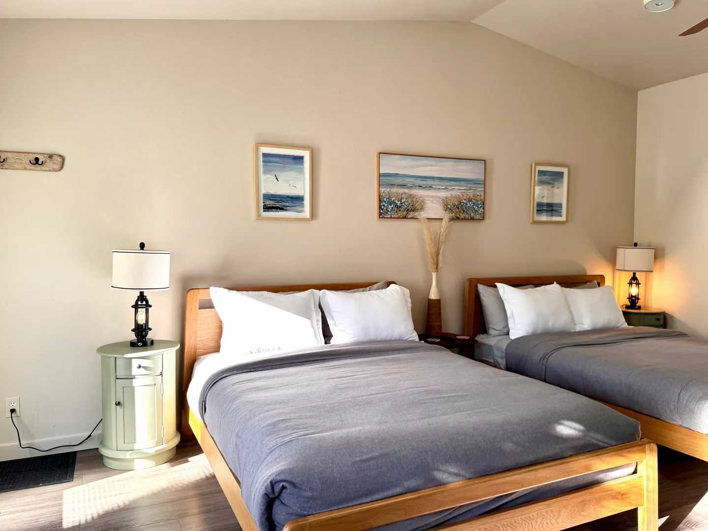 Bedroom featuring two wooden beds with grey covers, white pillows, nightstands with lamps, and framed coastal artwork.