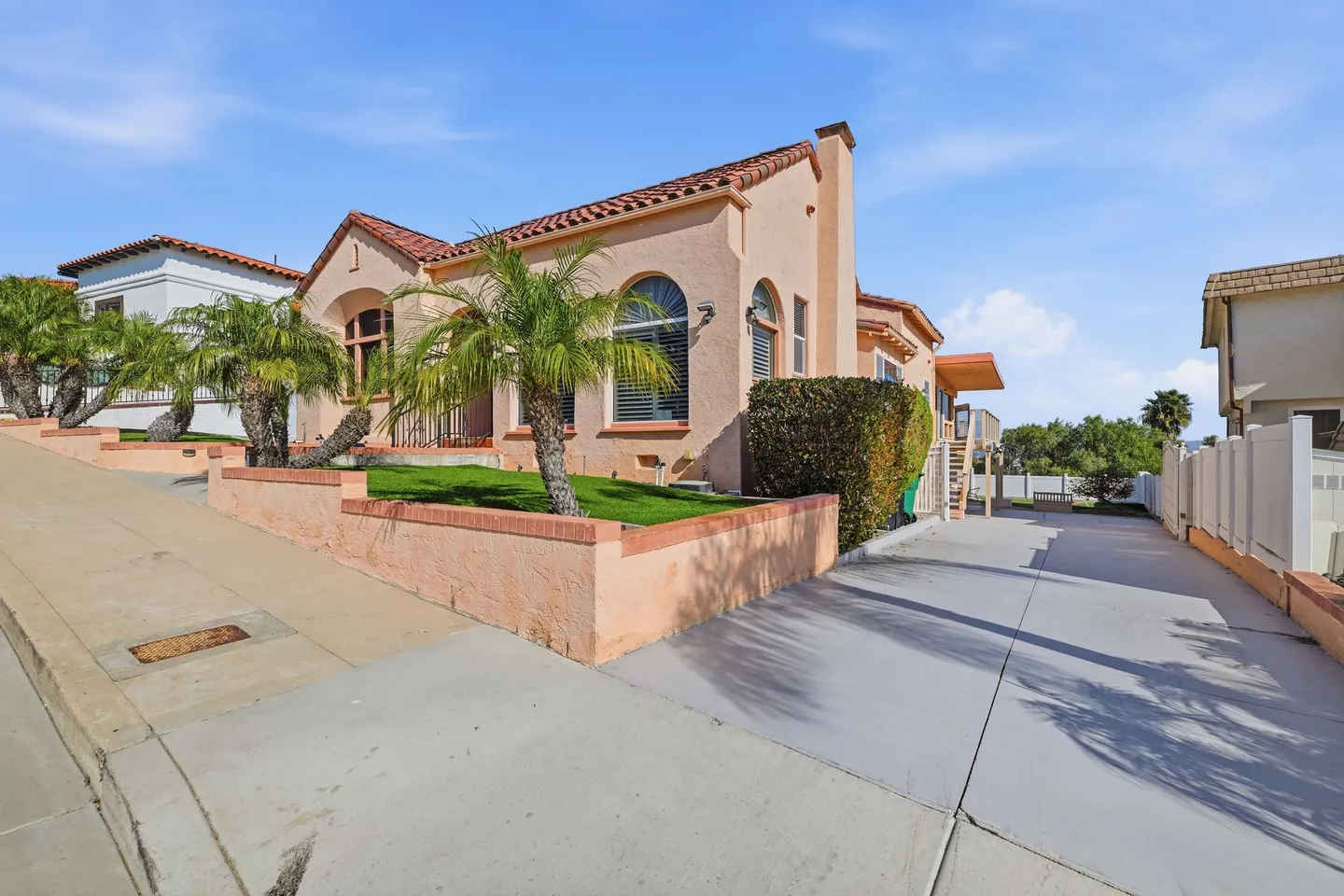 Exterior view of a peach-colored Mediterranean style home with a red tile roof, arched windows, and small palm trees along a sloped sidewalk.