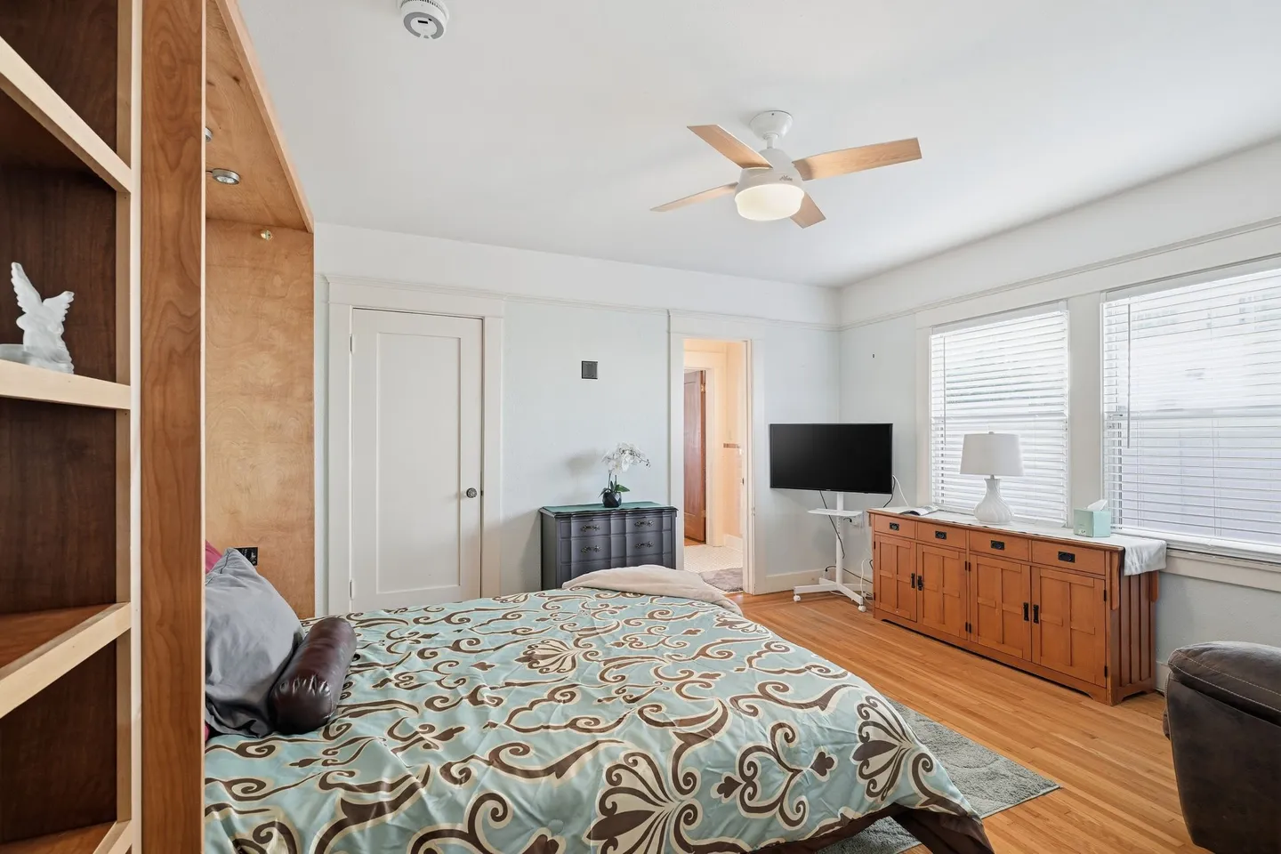 Bedroom with patterned bedding, a ceiling fan, a TV on a stand, wooden cabinets, and wood flooring.