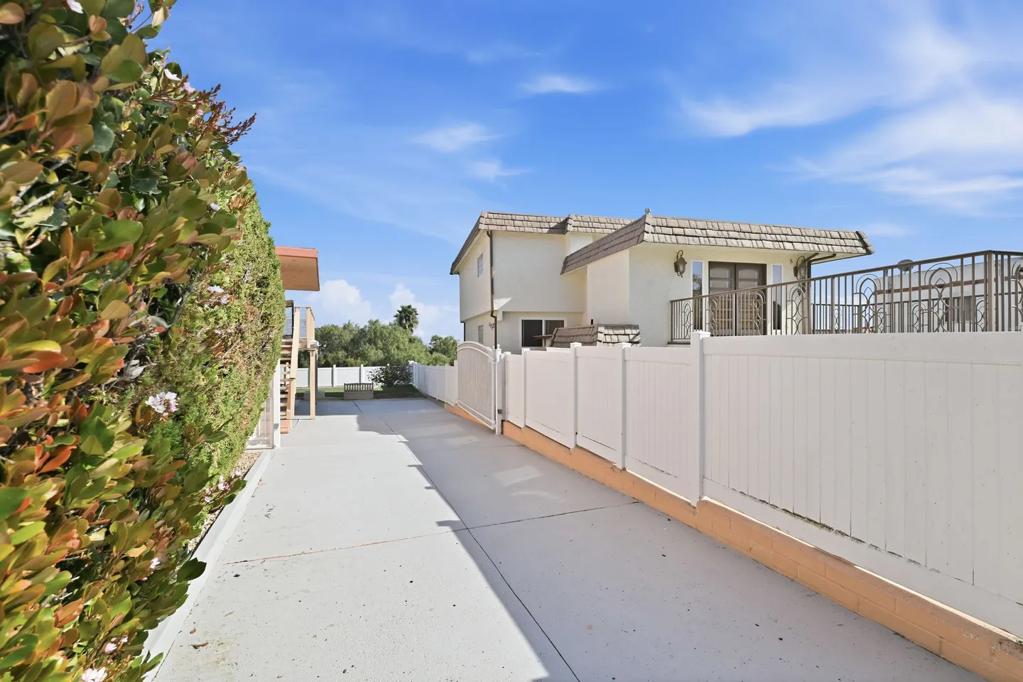 Concrete walkway bordered by a dense tall hedge on the left and a white fence on the right leading toward a two-story home under a blue sky.