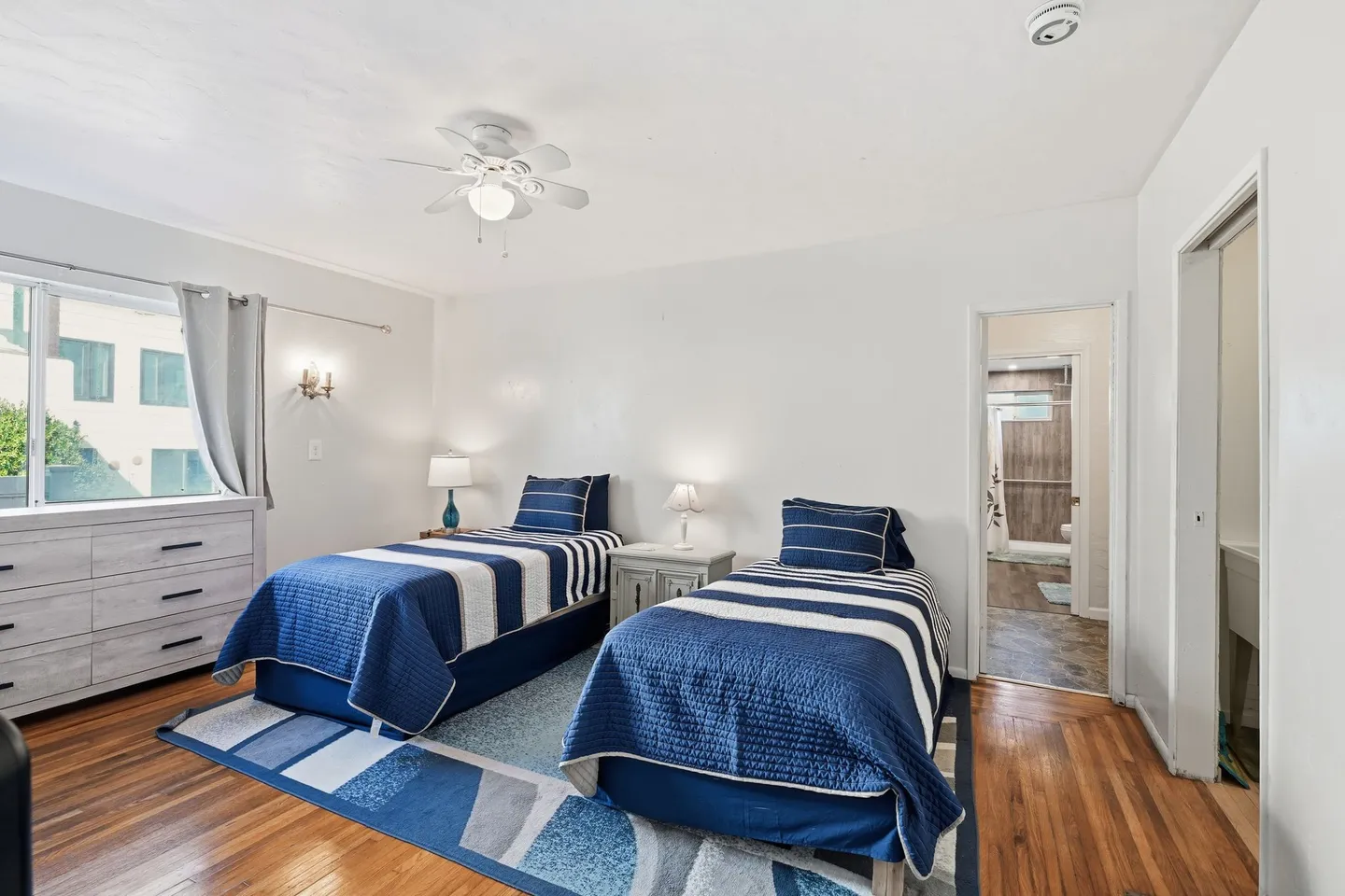 Bedroom with two twin beds dressed in blue and white bedding, hardwood floors, and an open doorway showing an en suite bathroom.