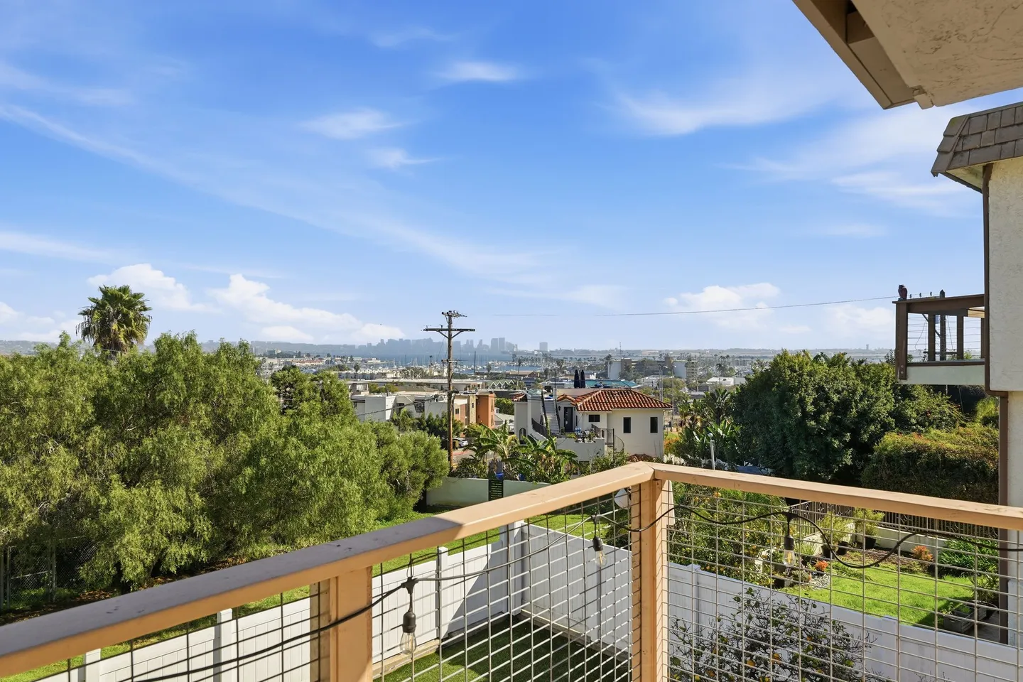 Elevated view from an outdoor balcony overlooking surrounding neighborhood houses, greenery, and a distant city skyline under a clear blue sky.