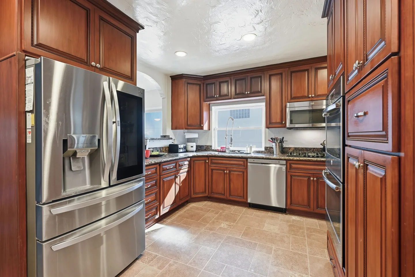 Kitchen featuring warm wood cabinets, tiled floors, granite countertops, and stainless steel appliances including a French-door refrigerator, double wall oven, dishwasher, and microwave.
