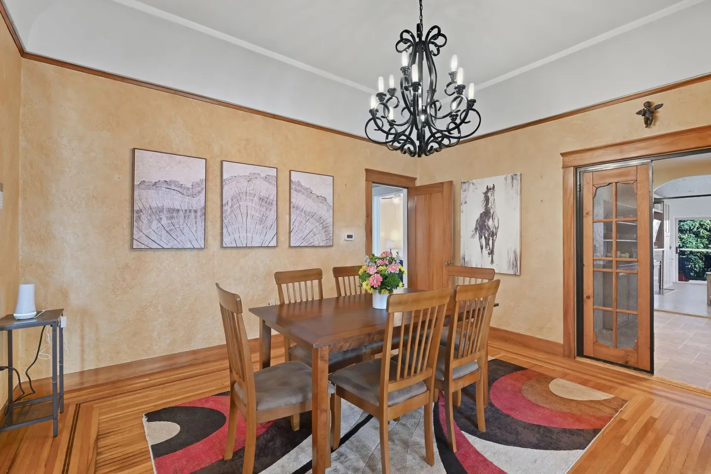 Dining room with a wooden table, chairs, an ornate black chandelier, patterned area rug, and artwork on textured beige walls.