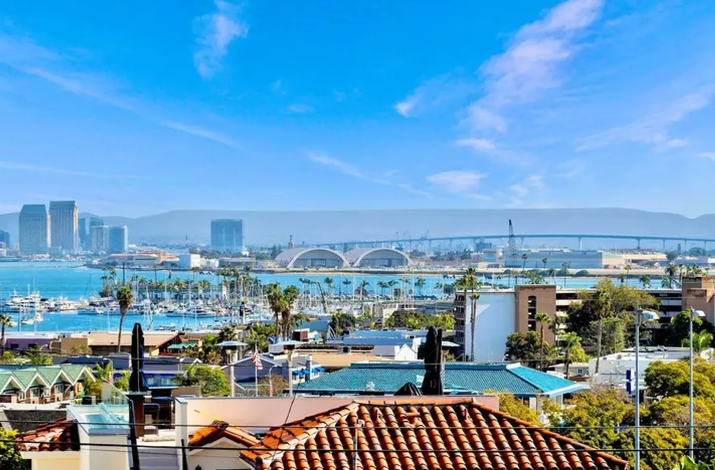 Elevated view overlooking rooftops toward a bay with boats, city skyline, and a large bridge under a bright blue sky.