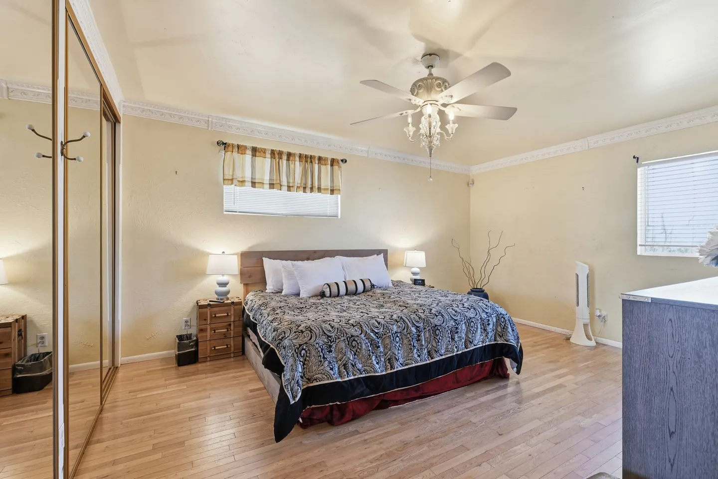 Bedroom featuring a made bed with an ornate patterned comforter, light wood flooring, mirrored closet doors, and a ceiling fan with a chandelier fixture.
