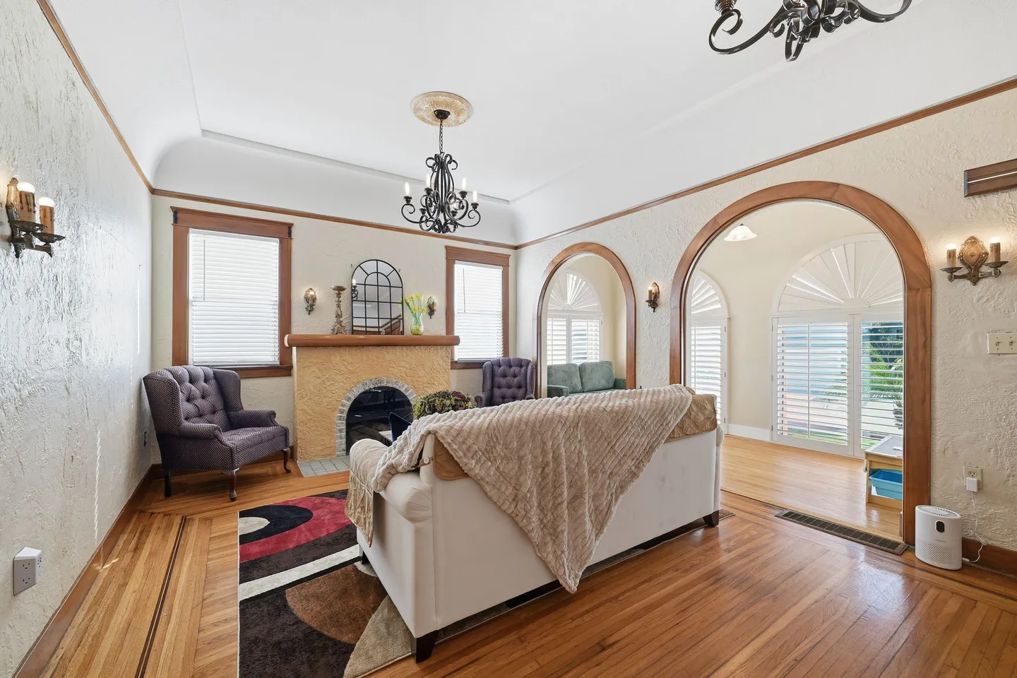 Living room with wood flooring, textured walls, a decorative fireplace, armchairs, and wood-trimmed arched doorways leading to an adjacent room.