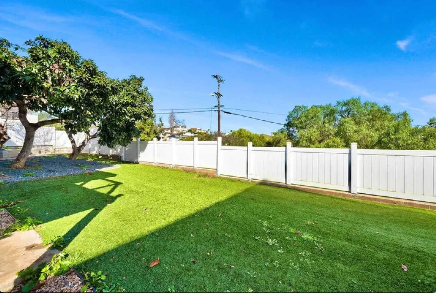 A fenced backyard featuring a green lawn, mature trees, and a white privacy fence under a clear blue sky.