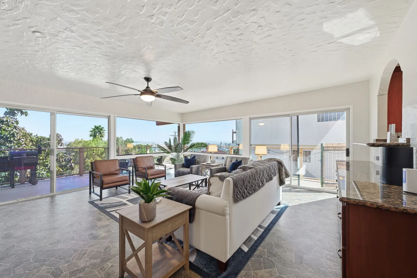 Sunroom or living area with textured tile floor, modern sofa and armchairs, ceiling fan, and large windows with sliding glass doors opening to an outdoor deck.