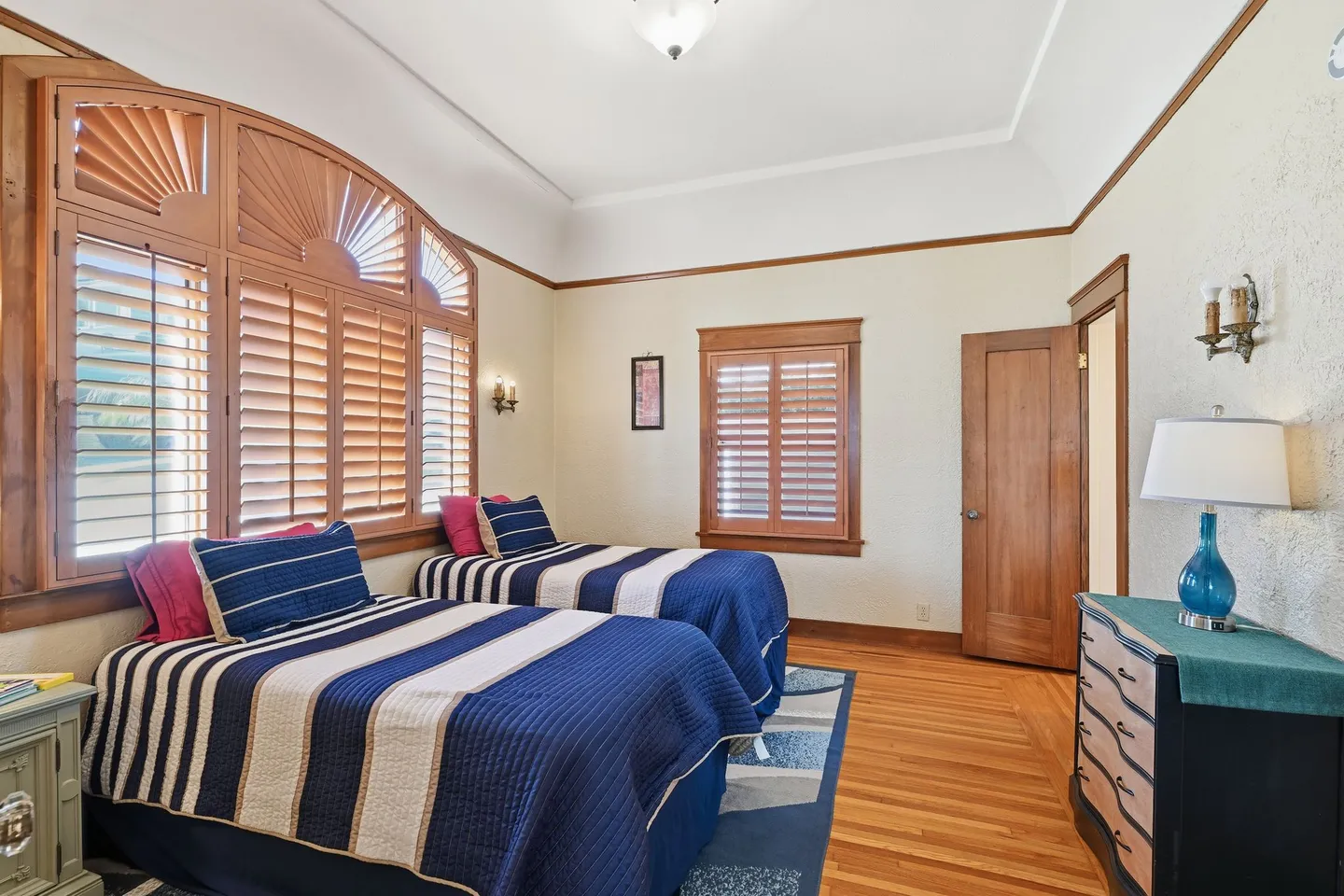 Bedroom featuring two twin beds with blue striped quilts, hardwood floors, and wooden plantation shutters covering arched and rectangular windows.