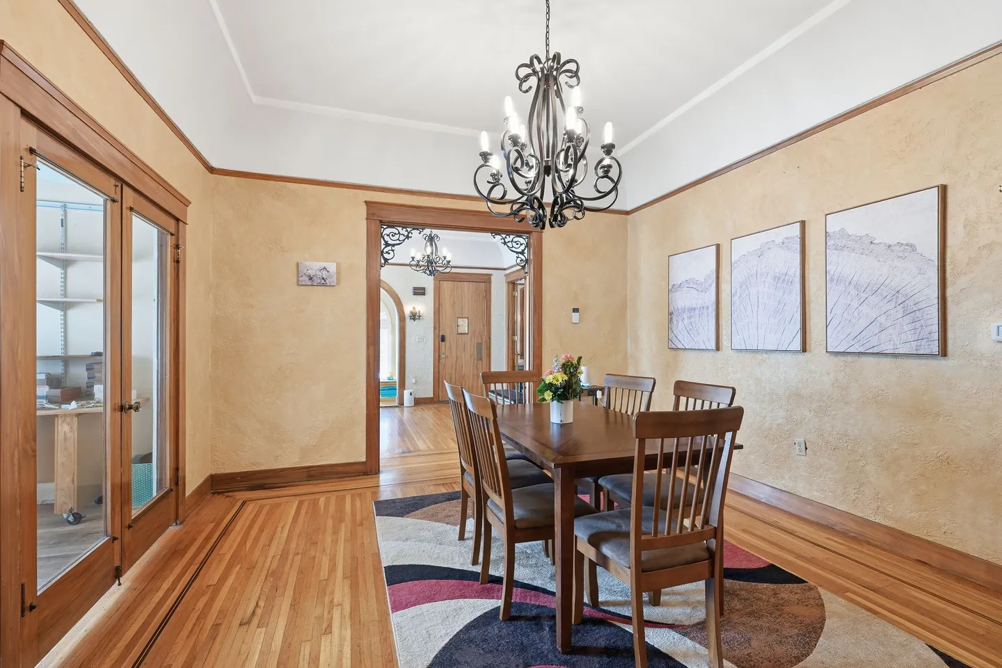 Dining room with a wooden table and chairs on a patterned area rug, lit by a black chandelier.