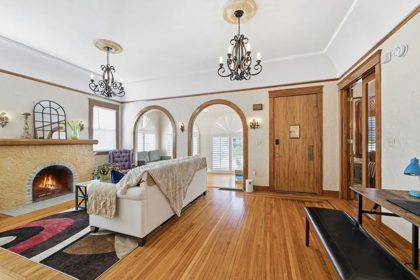 Living room with hardwood floors, an arched fireplace with a lit fire, black chandeliers, and arched doorways leading to an adjacent sunroom.