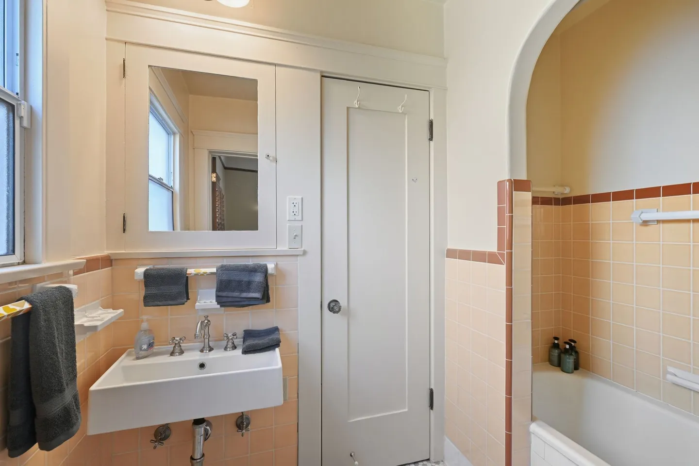 Bathroom with vintage peach tile, a wall-mounted sink, built-in medicine cabinet, and arched tub enclosure.