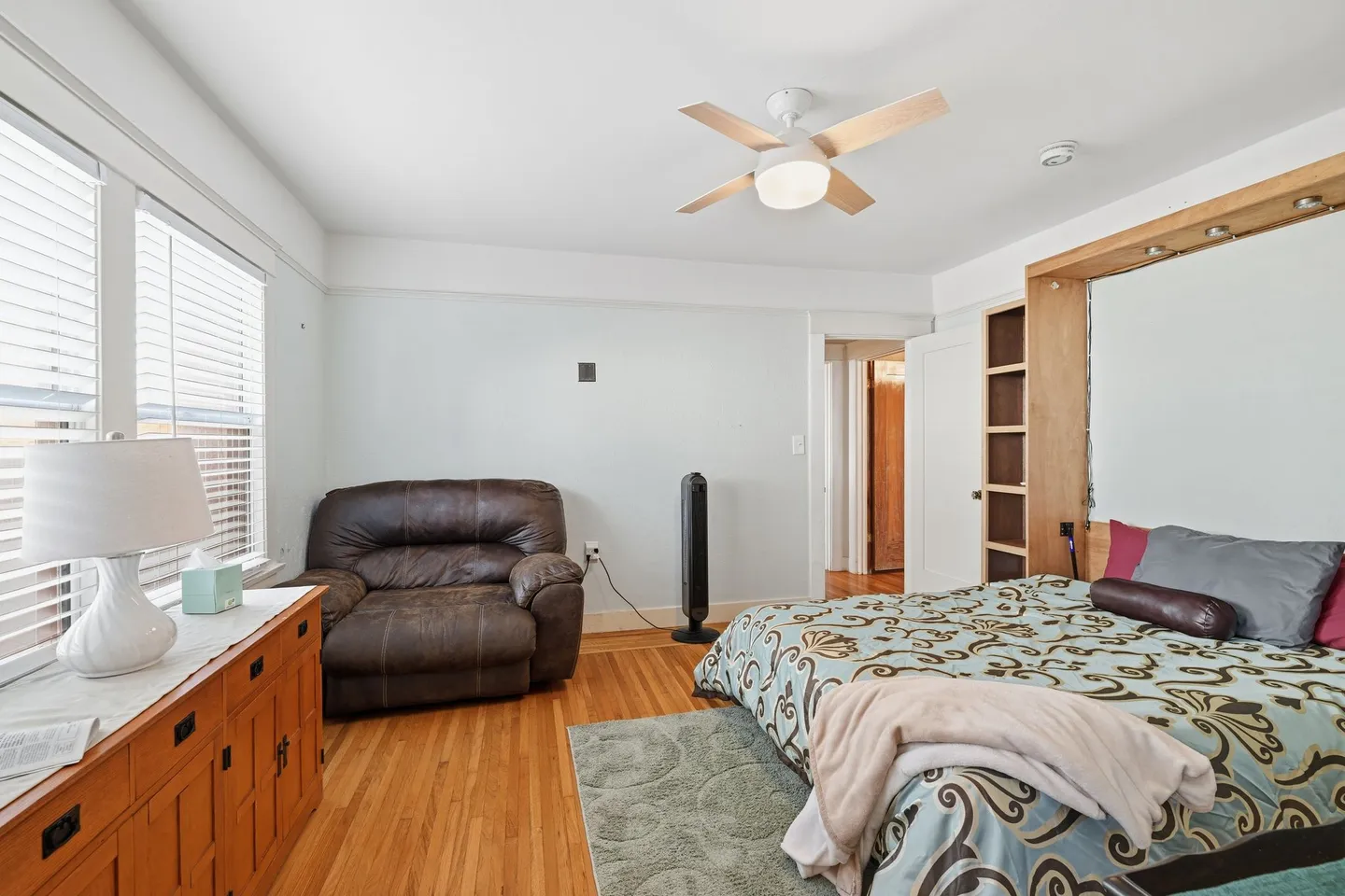 Bedroom with wood floors, patterned bedding, an oversized brown armchair, dresser, and built-in headboard unit.