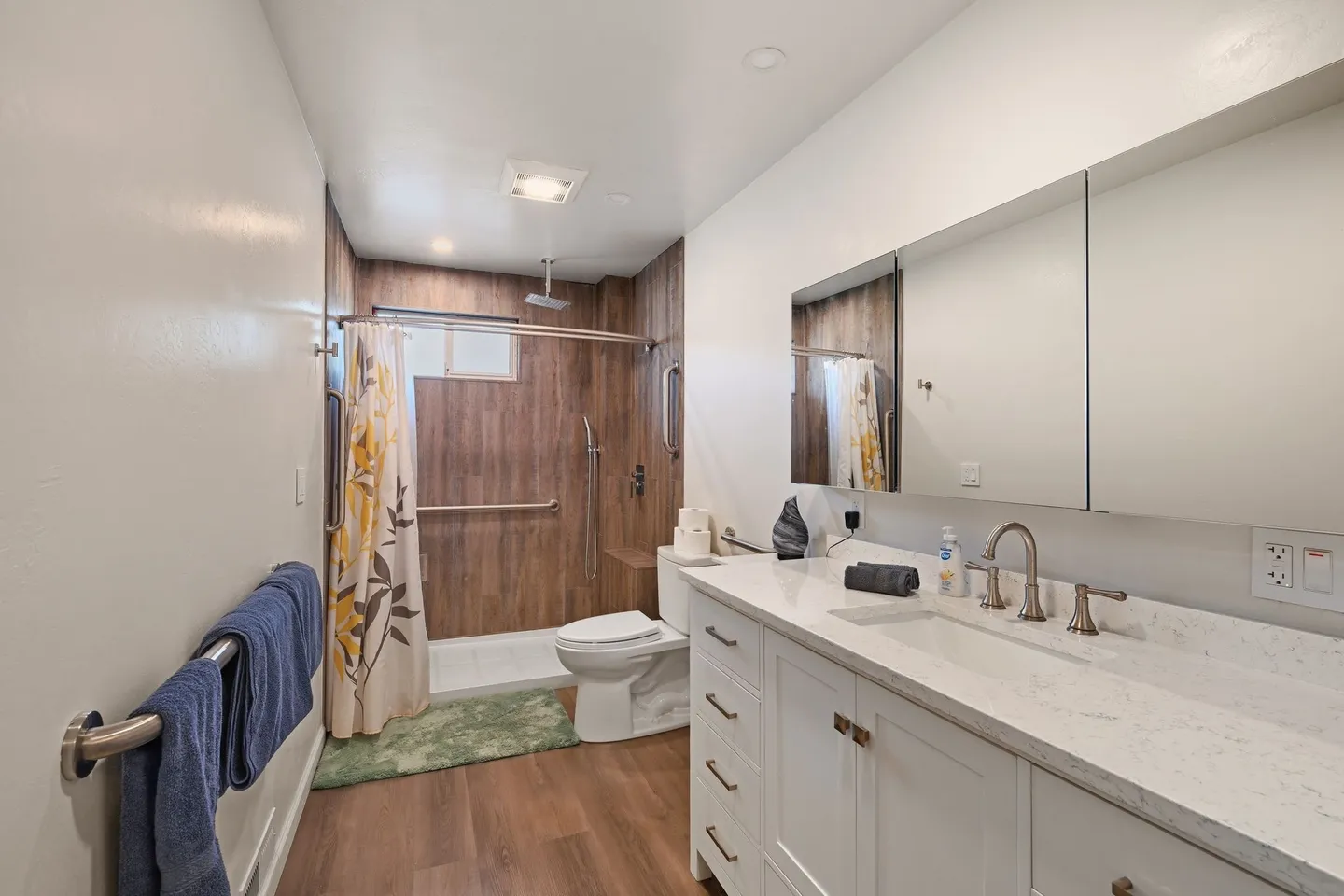 Bathroom featuring a white vanity with marble-patterned countertop, large mirrored medicine cabinets, a toilet, and a walk-in shower with wood-look tile.