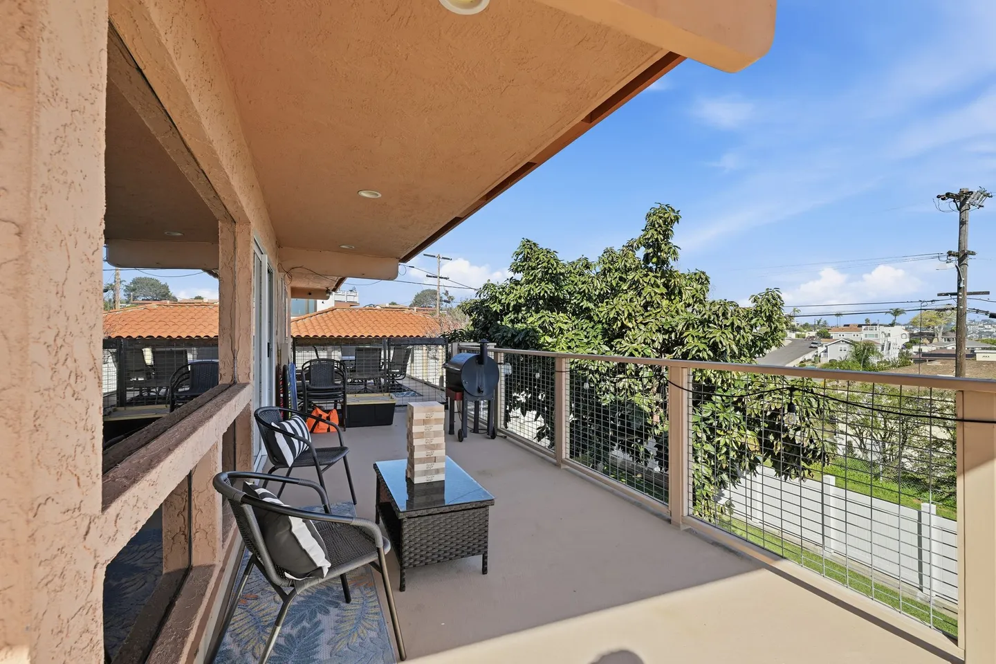 Elevated outdoor balcony with wire railing, outdoor chairs, small coffee table, and a barbecue smoker under a clear blue sky.