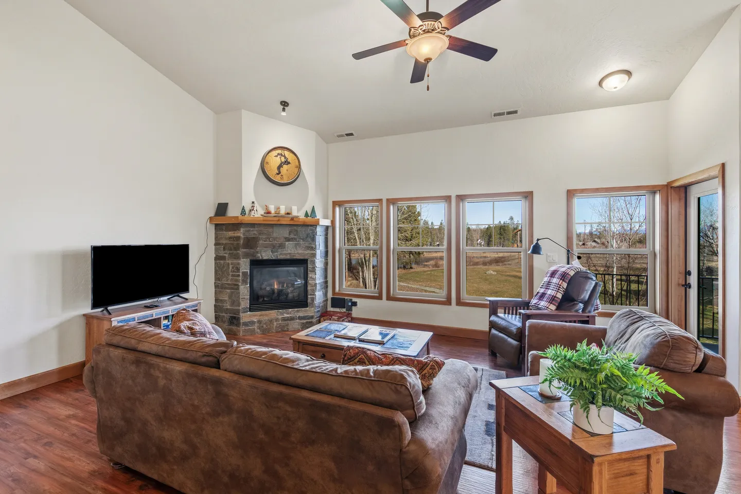 Living room with brown leather seating, a stone corner fireplace, flat-screen TV, and large windows overlooking trees.