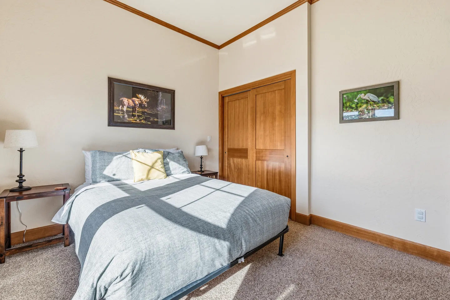 Bedroom with carpeted floor, made bed under framed artwork, wooden closet doors, and wood trim.