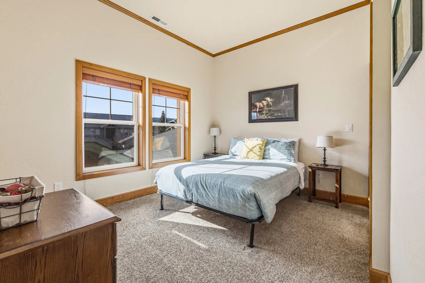 Bedroom with carpet flooring, a bed between two nightstands, and two windows with wood trim showing daylight.