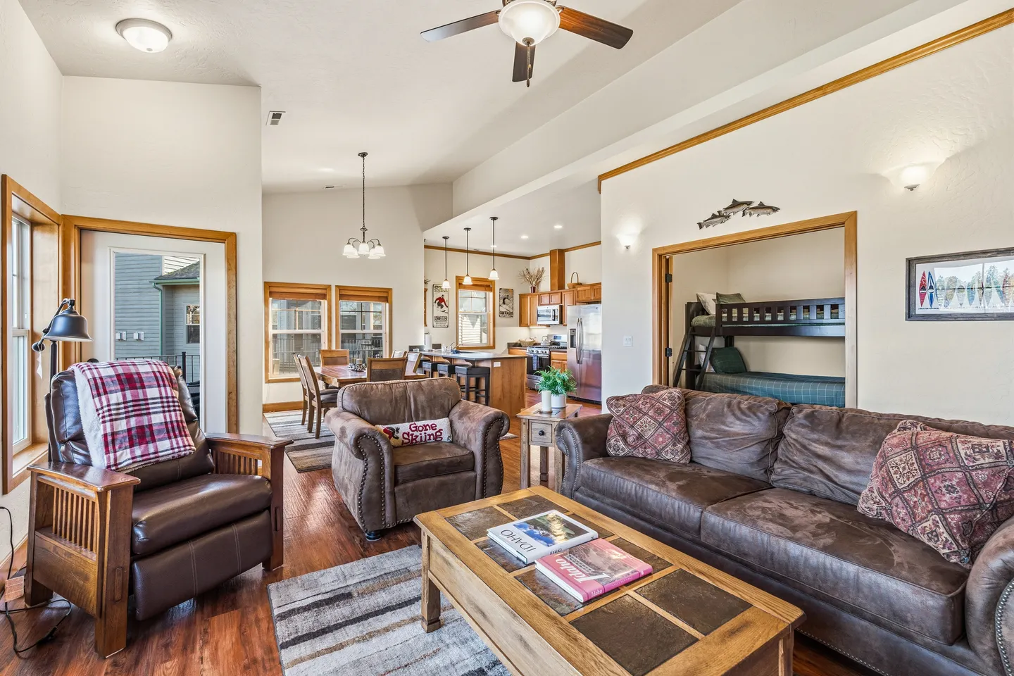 Open-concept living room with leather seating, dining area, kitchen, and an adjacent bunk room visible through a wide doorway.