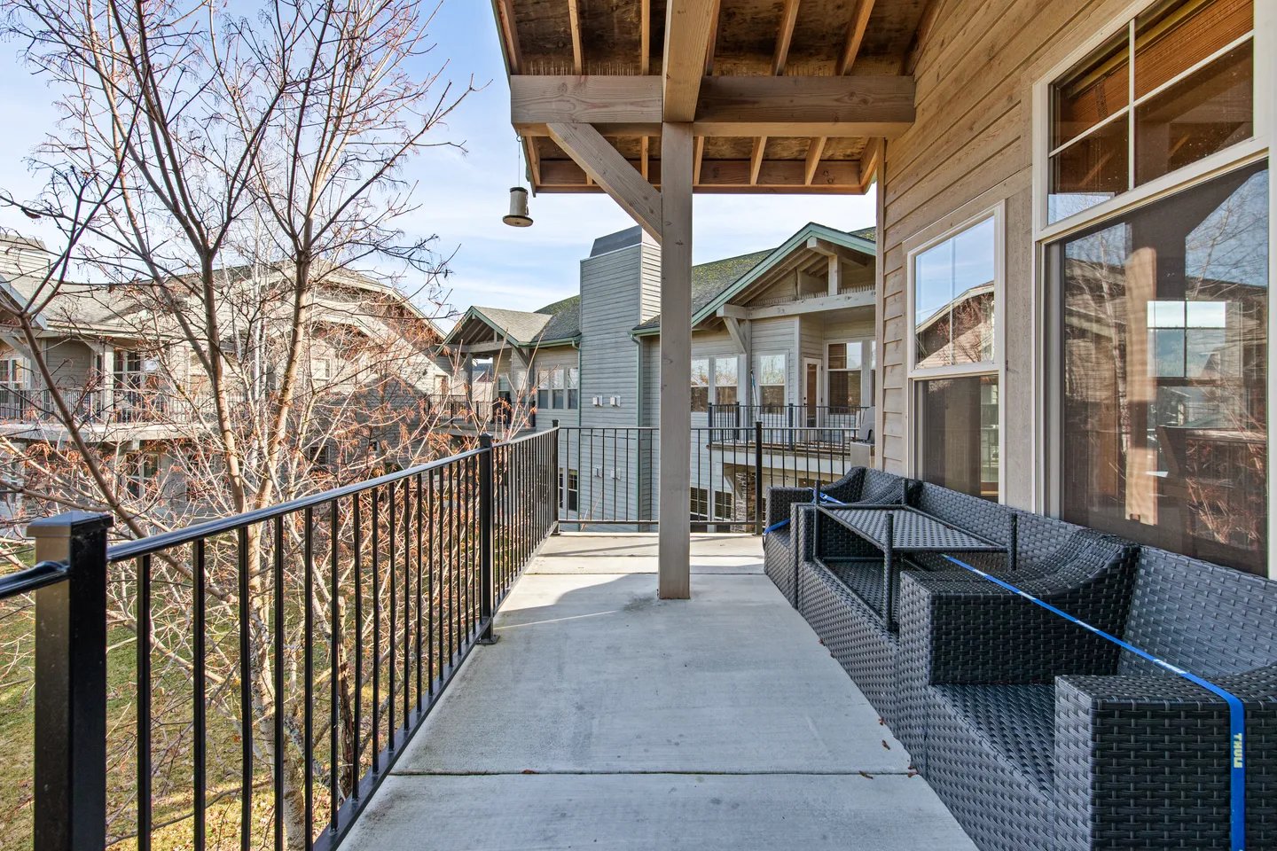 A covered concrete balcony with black metal railings, outdoor wicker seating, and views of nearby buildings and trees.