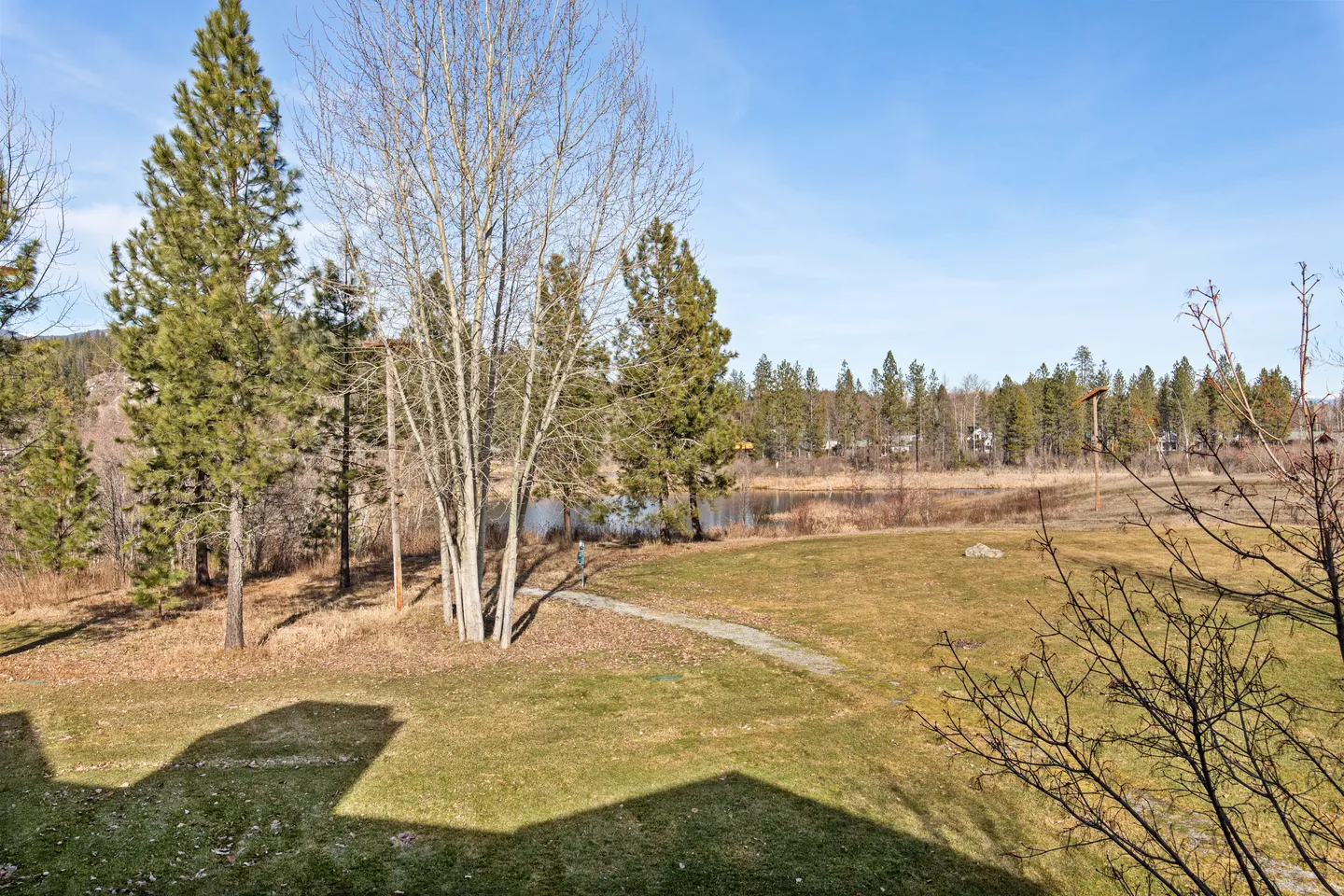 Elevated outdoor view of a grassy yard with trees, a walking path, and a pond in the distance under a blue sky.