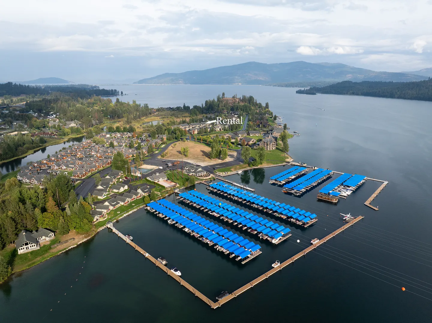 Aerial view of a lakeside marina with covered boat slips next to a residential community, with distant mountains and water.