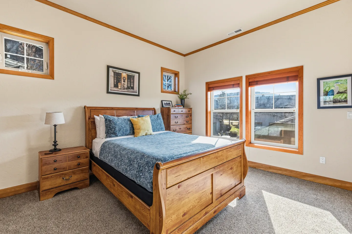 Bedroom featuring a wooden sleigh bed with blue and yellow bedding, matching wooden nightstand and dresser, carpet, and multiple windows with wood trim.