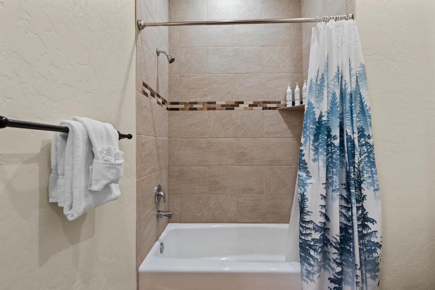 Bathroom bathtub and shower combination featuring tiled walls with a mosaic accent strip, toiletries on a corner shelf, and a pine tree pattern shower curtain.