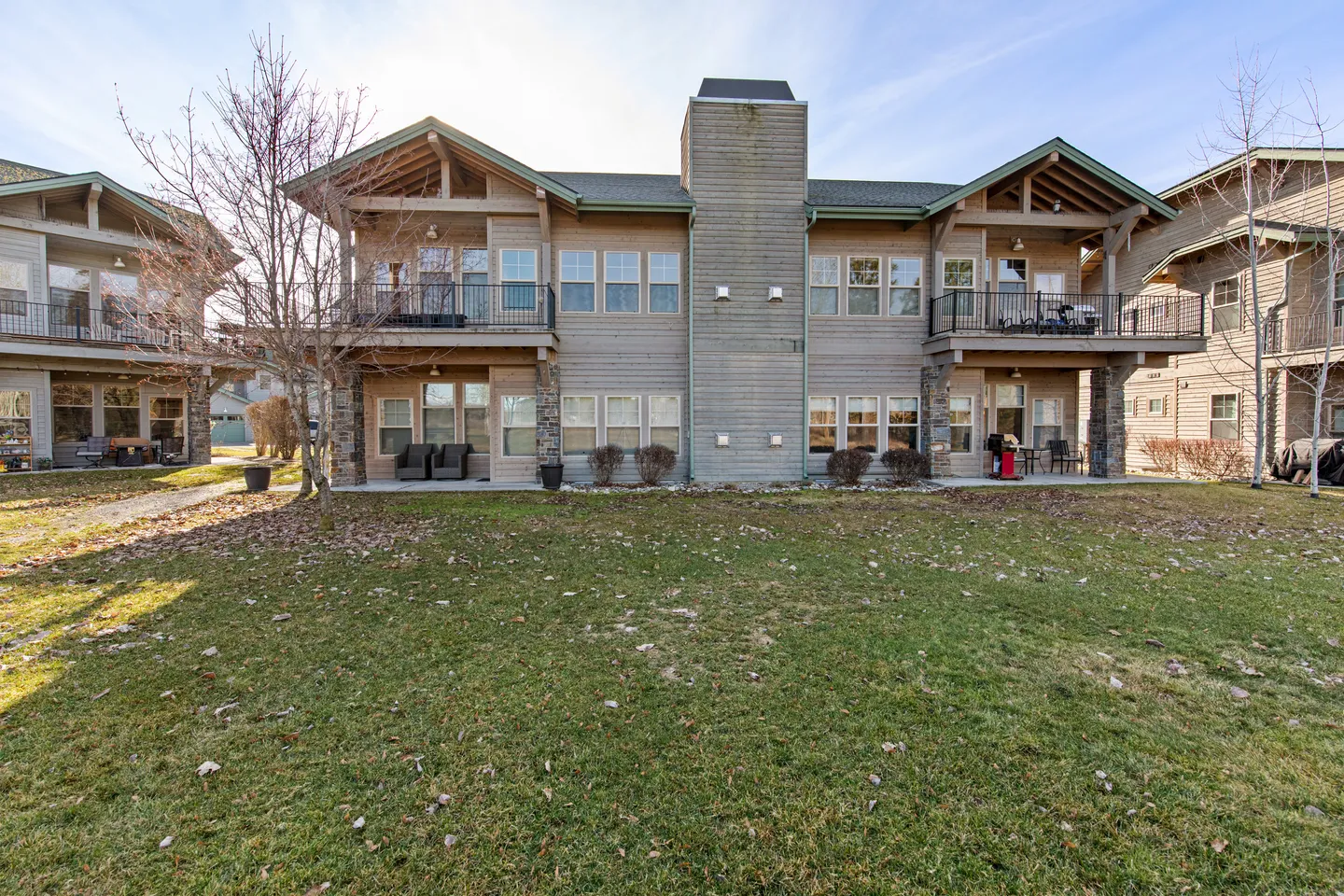 Exterior view of a multi-unit two-story residential building with wooden siding, balconies, ground-floor patios, and a grassy lawn in the foreground.