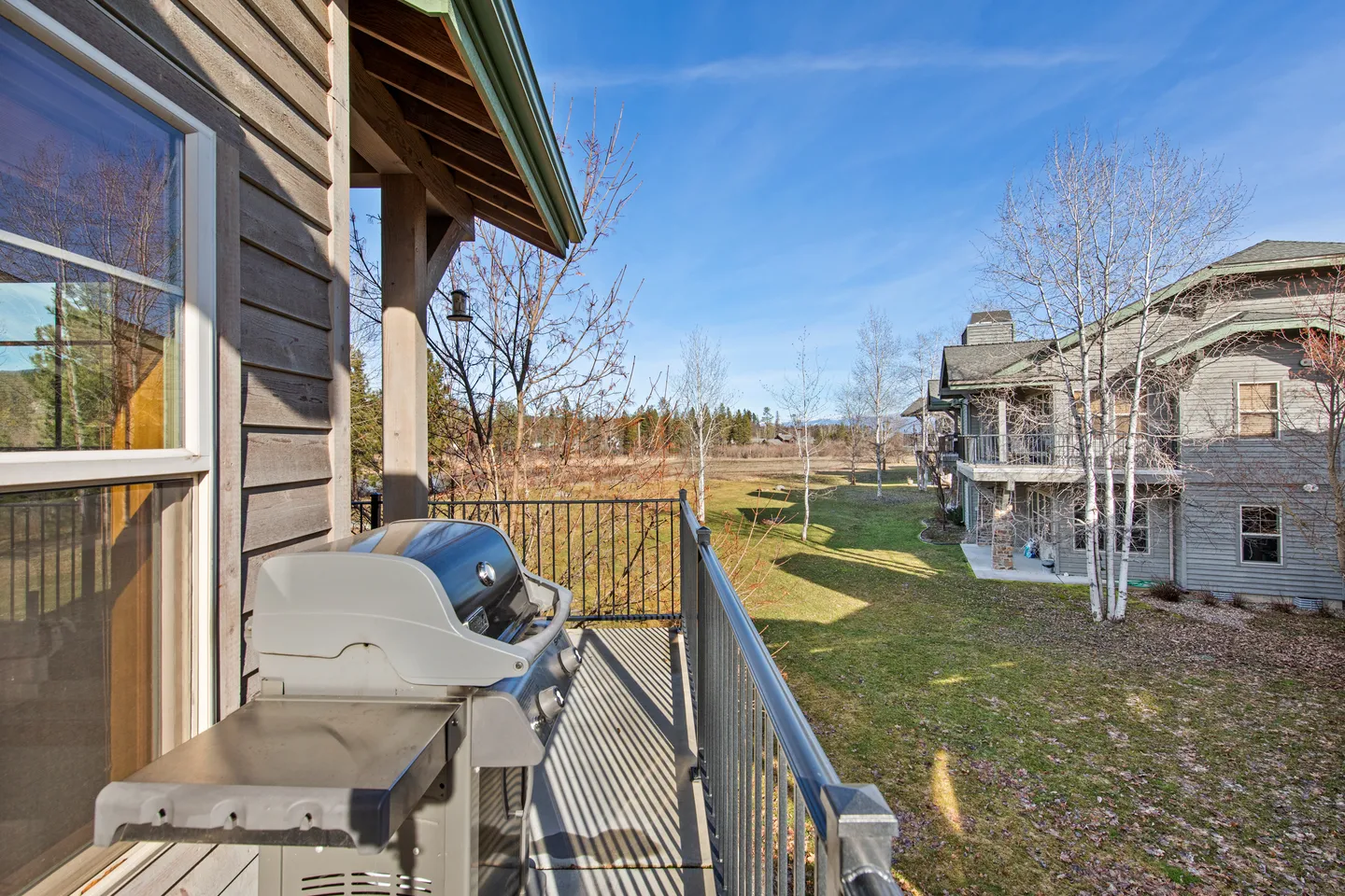 Balcony with a barbecue grill overlooking a grassy yard, nearby trees, and neighboring residential buildings under a blue sky.