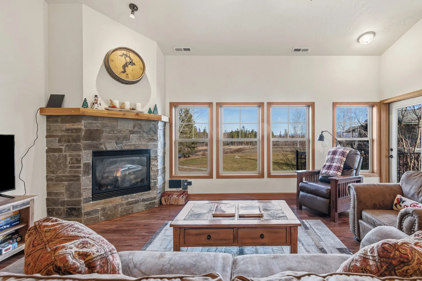 Living room with a stone fireplace, wood-framed windows looking out onto a landscape, coffee table, and comfortable seating.