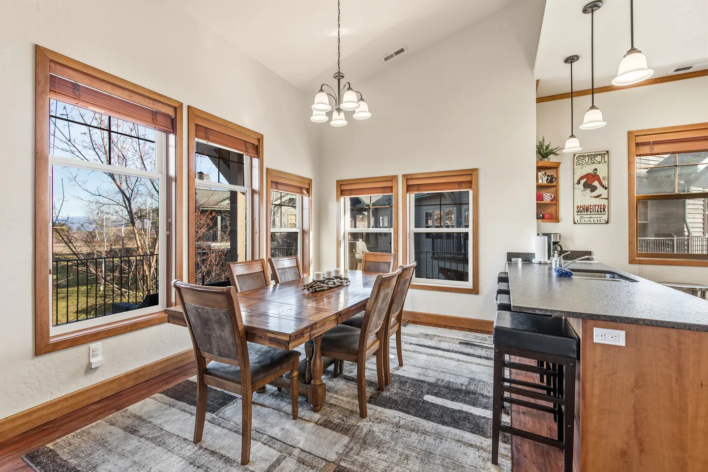 Dining area with a wooden table, chairs, an area rug, and wood-trimmed windows adjacent to a kitchen counter.