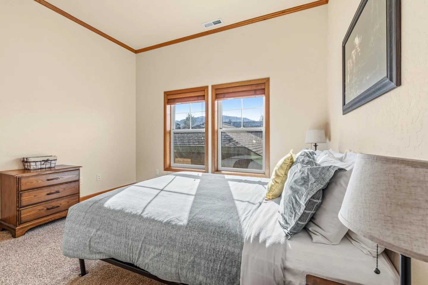 Bedroom with carpet, a made bed, two wood-trimmed windows with views of distant mountains, and a wooden dresser.