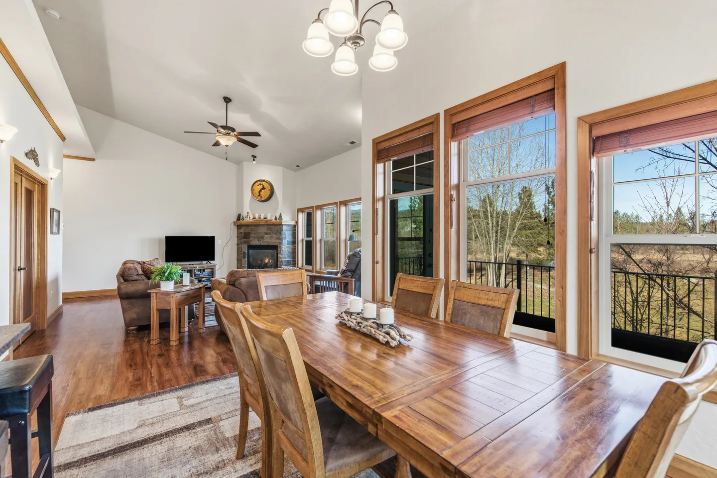 Dining area with a wooden table and chairs, large windows, and a view into the adjacent living room featuring a stone fireplace.