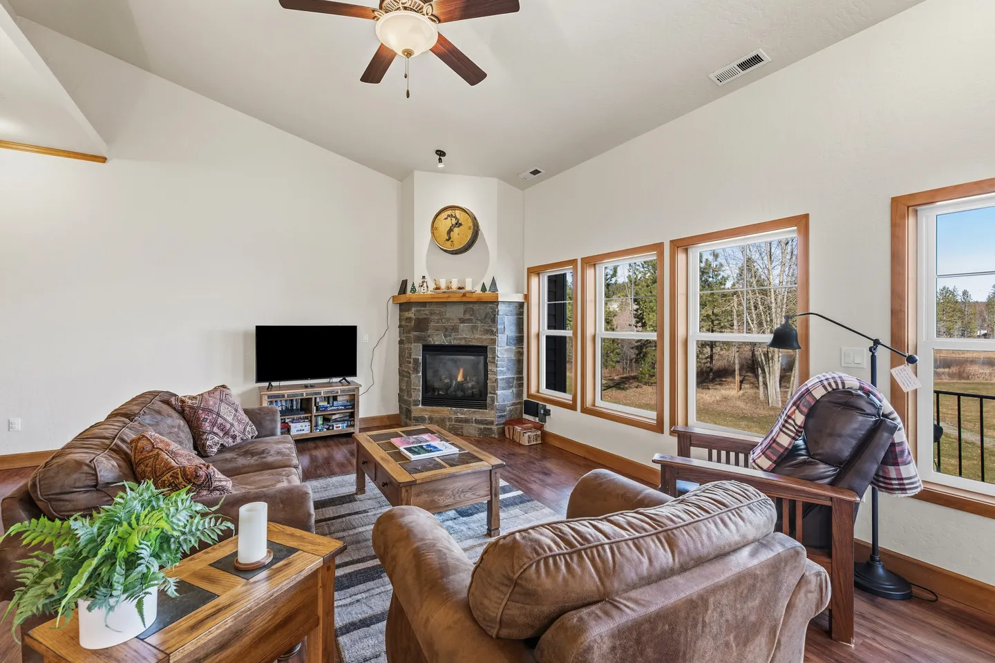 Living room with brown leather seating, a stone corner fireplace, television, coffee table, and large windows looking outside.