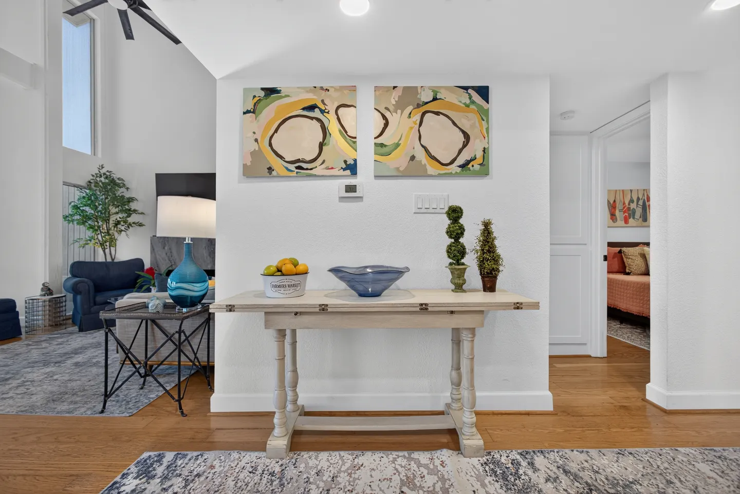 A wooden console table decorated with a fruit bucket, glass bowl, and topiaries beneath abstract art in an open hallway.