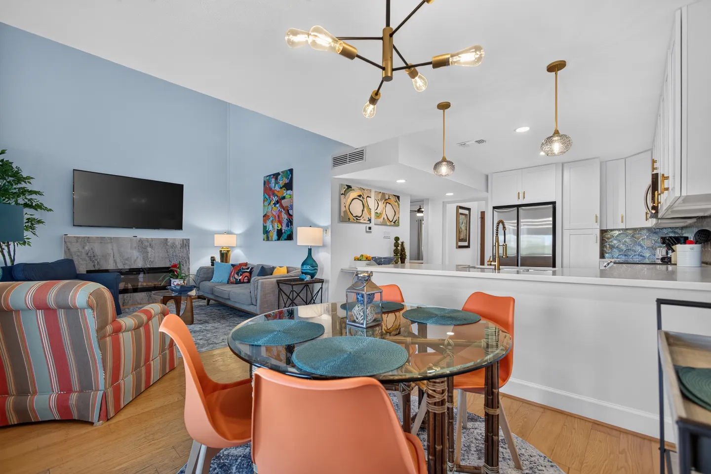 Open-concept dining, living room, and kitchen featuring a glass dining table with orange chairs, modern light fixtures, and a fireplace with a mounted TV.