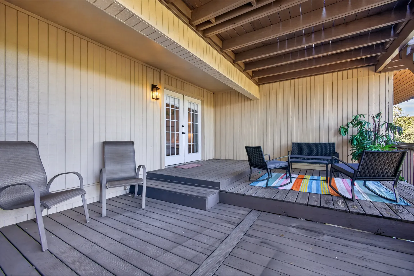 Covered wooden deck with outdoor seating, area rug, and double doors leading into the home.