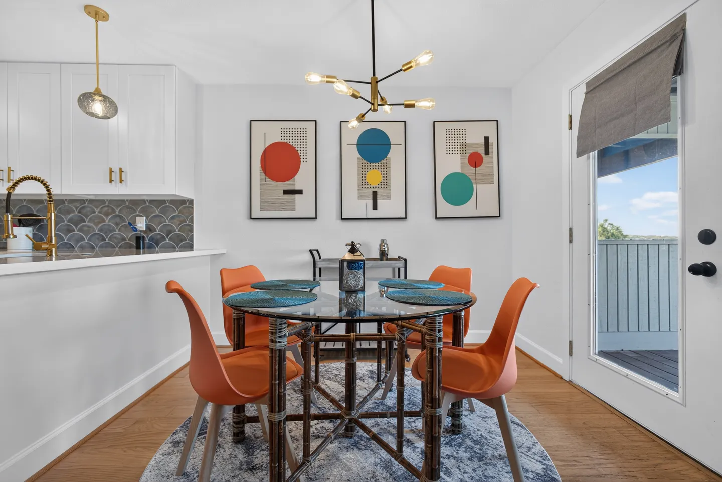 Dining area with a round glass-top table, four orange chairs, modern chandelier, and kitchen breakfast bar to the left.